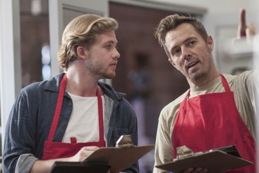 Zwei Mitarbeiter in roten Schürzen halten Klemmbretter und sprechen in einem Geschäft miteinander.